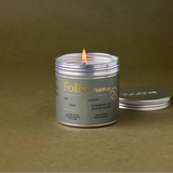 A lit silver metal tin candle with a wick, shown on a textured olive green background with its matching lid resting beside it. The label reads 'Folk by FieldDay'.