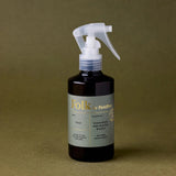 A dark amber spray bottle with a white trigger sprayer and a muted sage-green label reading 'folk by Fiebo' and 'Evergreen and Woody Wood', set against a solid olive-brown background.