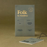 A square fragrance tag for 'Folk by FieldDay' stands upright on a textured brown surface, with a smaller matching tag placed in front and a black string attached. The packaging is a muted gray-green with gold typography and a small circular logo reading 'Folk of the North'.